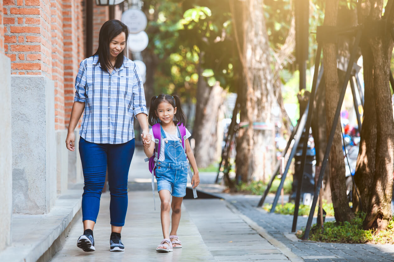 An adult and a young girl wearing a backpack walk hand in hand on a tree-lined sidewalk in the daytime.