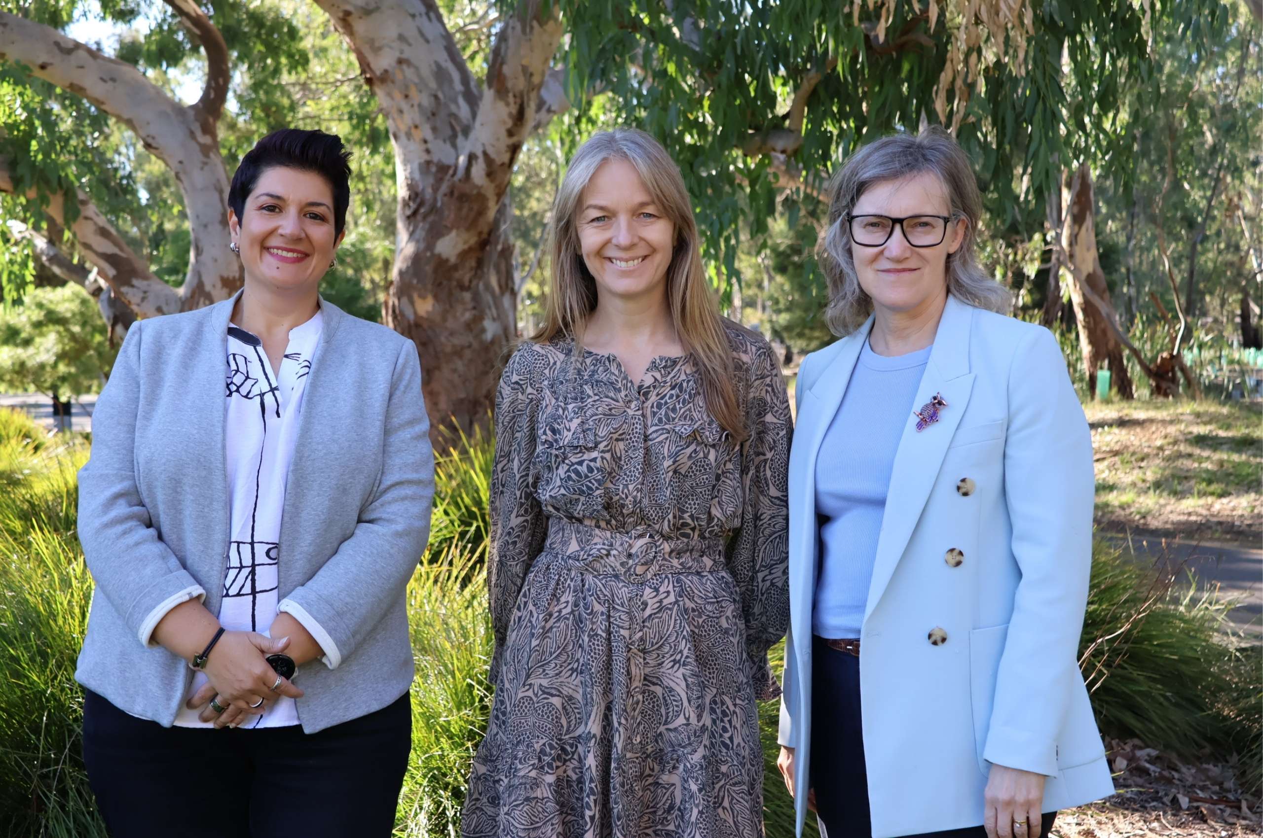Three women in business dress smiling at the camera. A lush Australian garden in the background