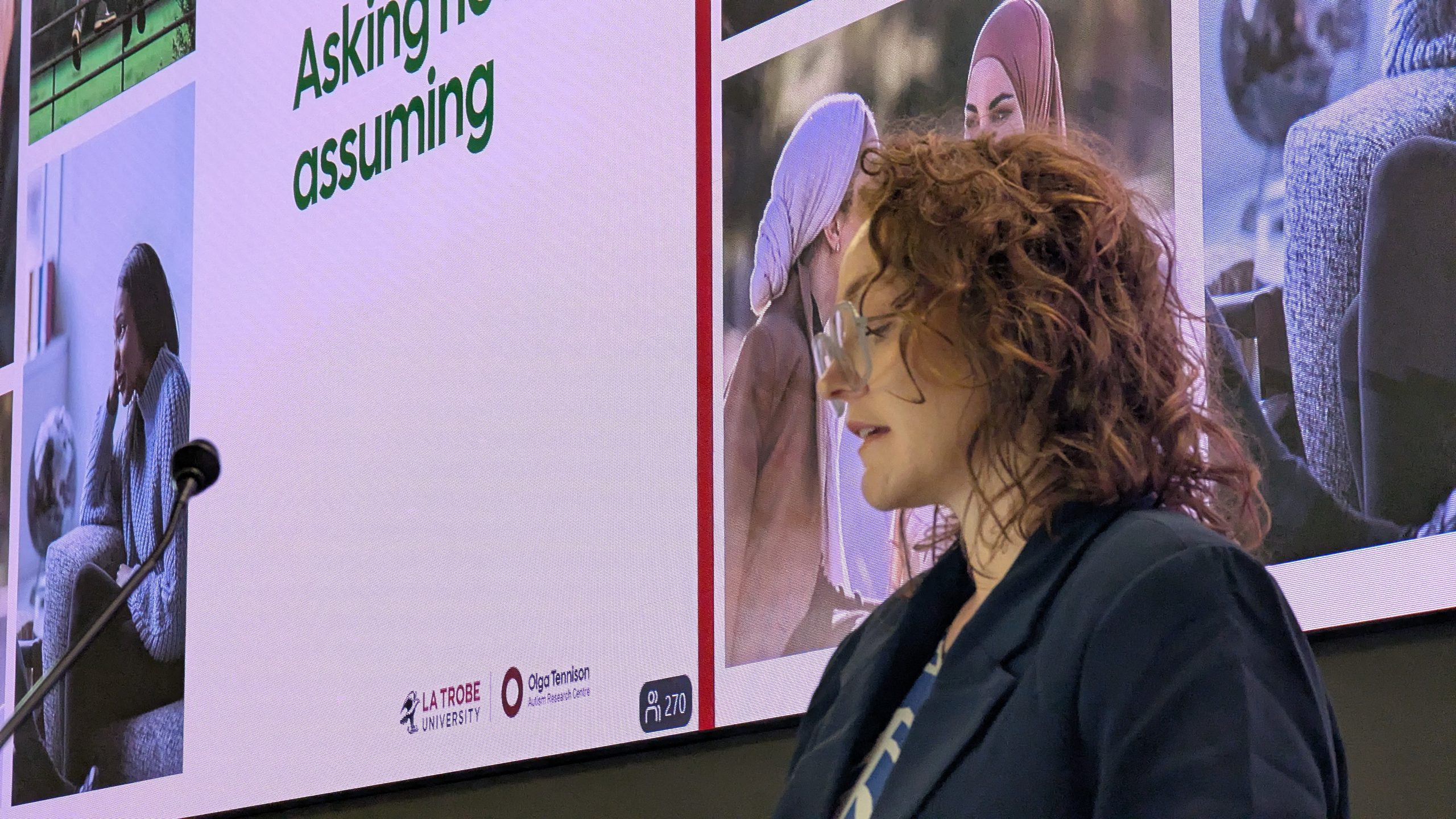 A person with curly reddish-brown hair and glasses is shown in profile, standing in front of a large presentation screen at an event. The screen displays photographs of people participating in a discussion or presentation, with visible La Trobe University and Olga Tennison Autism Research Centre logos and a slide heading beginning “Asking not assuming”.